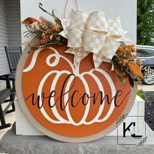 May include: A wooden pumpkin-shaped door hanger with the word "welcome" written in white script. The pumpkin is painted orange and has a white stem. The hanger is decorated with a white and beige checkered bow and fall foliage.