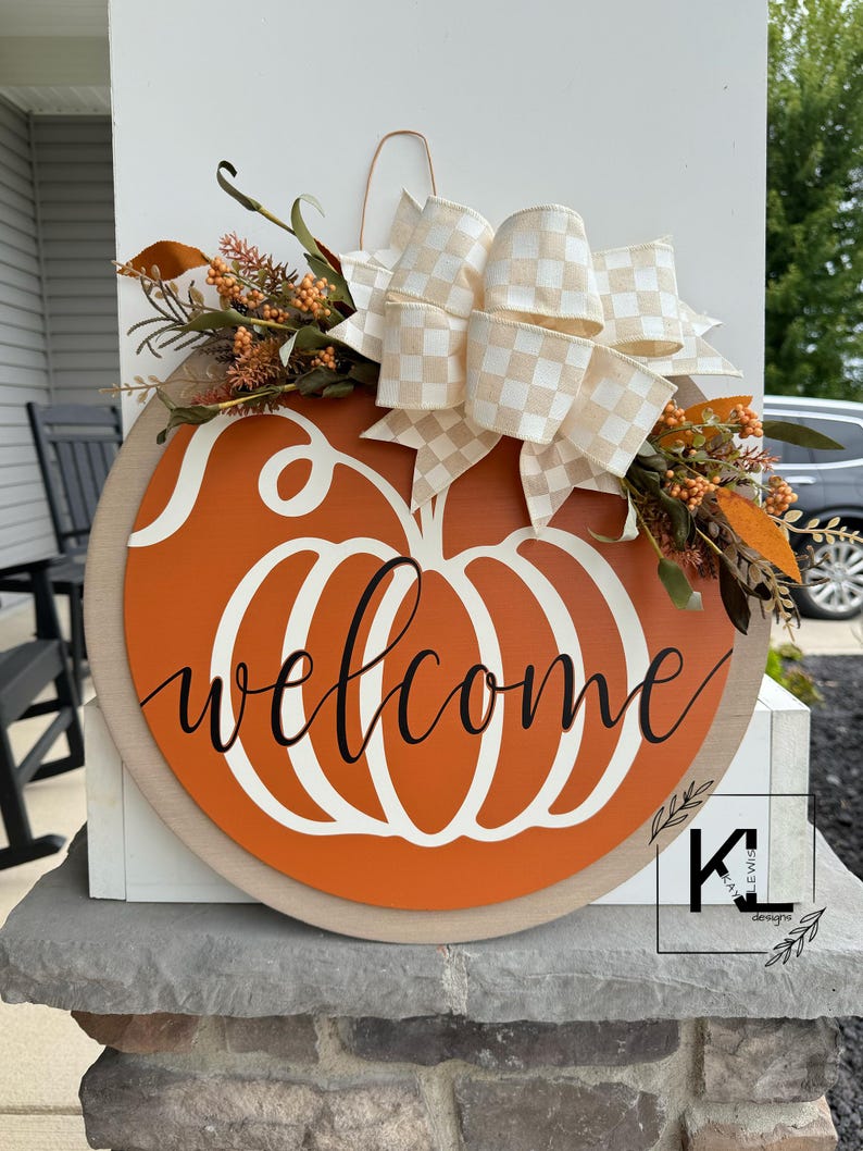 May include: A wooden pumpkin-shaped door hanger with the word "welcome" written in white script. The pumpkin is painted orange and has a white stem. The hanger is decorated with a white and beige checkered bow and fall foliage.