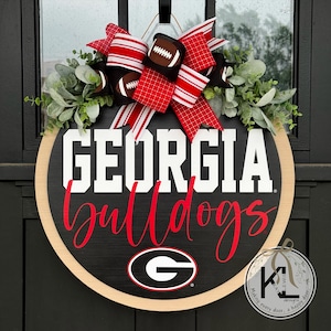 May include: A round wooden door wreath featuring the words "GEORGIA Bulldogs" in white and red lettering. The wreath has a black background with the Georgia Bulldogs logo at the bottom. It is decorated with a red and white striped bow, football accents, and greenery.