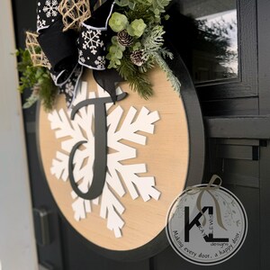 May include: A wooden round door hanger with a black border. The center of the hanger features a white snowflake with a black letter "J" in the center. A black and white bow with snowflakes is attached to the top of the hanger. The hanger is decorated with greenery and pine cones. The text "Making every door, a happy door." is printed on a small circle with a black and white logo.