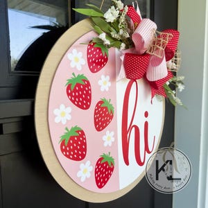 May include: A round wooden door hanger with a pink and white background. The pink side features a pattern of strawberries and white daisies. The white side says "hi" in red cursive. A red and white gingham bow with white polka dots is attached to the top of the hanger.