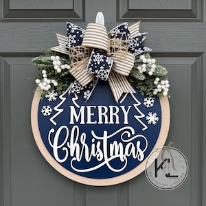 May include: A round wooden door sign with a dark blue center featuring the white text Merry Christmas and white snowflake accents. It is topped with a large bow made of navy blue snowflake ribbon and beige striped ribbon, plus white berry sprigs.