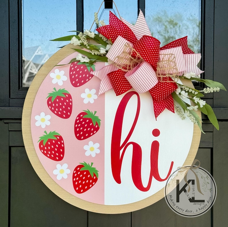 May include: A round wooden door hanger with a pink and white background. The pink side features a pattern of red strawberries and white daisies. The white side says "hi" in red cursive lettering. A red and white polka dot bow with a burlap center is attached to the top of the door hanger.
