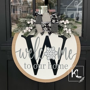May include: A round wooden door hanger with a white background and a black letter W. The letter W has a gray snowflake in the center. The words "welcome to our home" are written in gray cursive lettering. The door hanger is decorated with a black and white plaid bow and a sprig of faux greenery with white berries.