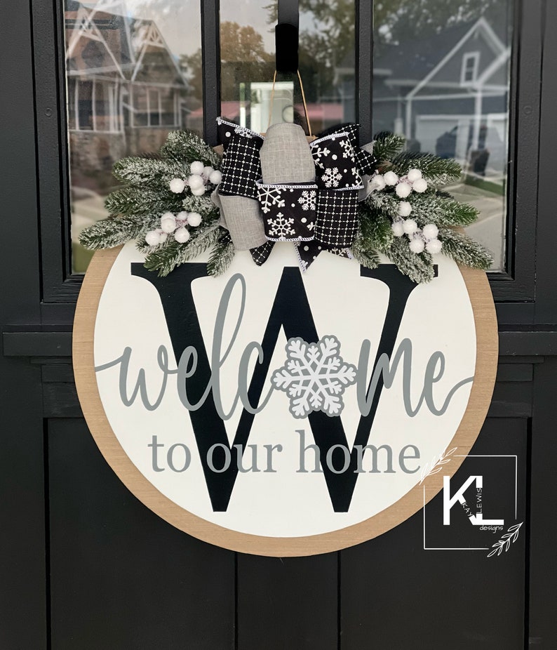 May include: A round wooden door hanger with a white background and a black letter W. The letter W has a gray snowflake in the center. The words "welcome to our home" are written in gray cursive lettering. The door hanger is decorated with a black and white plaid bow and a sprig of faux greenery with white berries.