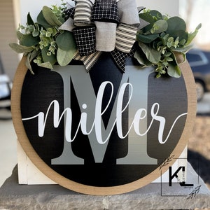 May include: A black and gray round wooden door hanger with the name "miller" written in white cursive script. The letter "M" is in gray and is larger than the rest of the name. The door hanger is decorated with a black and white striped bow and a wreath of green leaves.