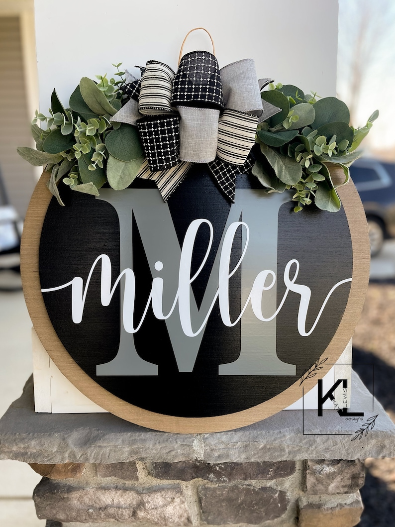 May include: A black and gray round wooden door hanger with the name "miller" written in white cursive script. The letter "M" is in gray and is larger than the rest of the name. The door hanger is decorated with a black and white striped bow and a wreath of green leaves.