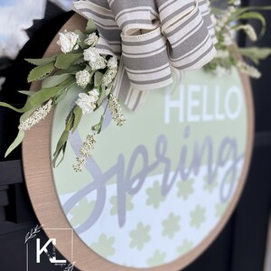 May include: A round wooden sign with a gray and white striped bow, white flowers, and the text "HELLO SPRING" in gray lettering. The sign has a green and white floral pattern.