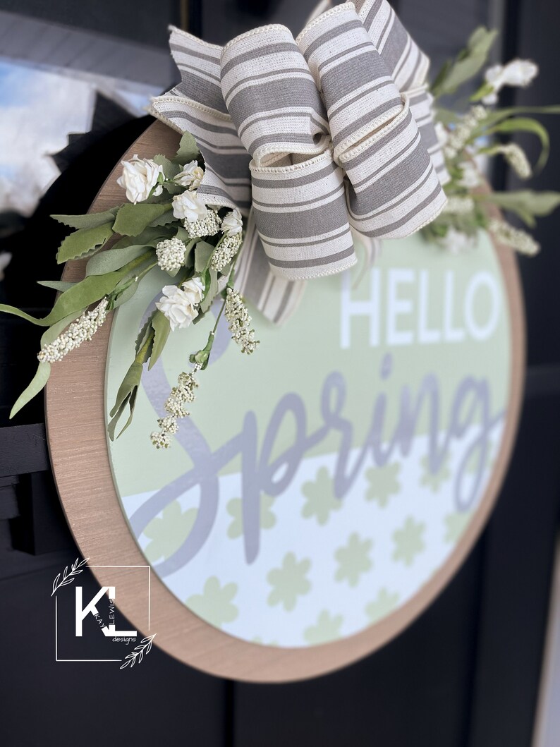 May include: A round wooden sign with a gray and white striped bow, white flowers, and the text "HELLO SPRING" in gray lettering. The sign has a green and white floral pattern.
