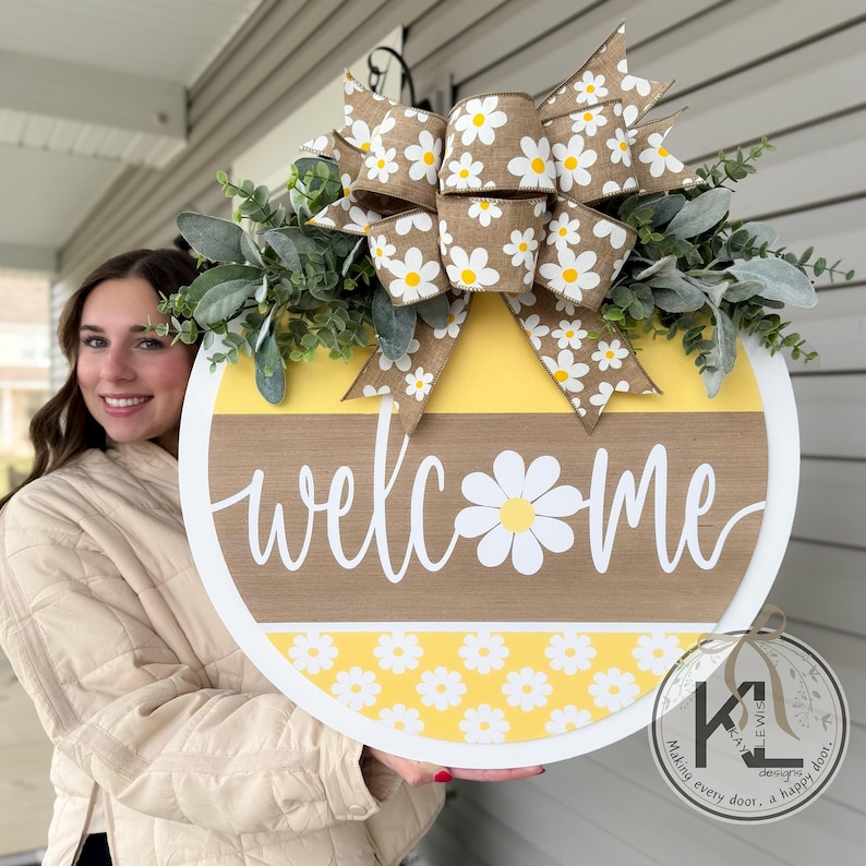 Op de afbeelding: Een ronde houten welkomstbord met een witte rand. Het bord heeft een gele en bruine achtergrond met het woord "welcome" in witte letters. Een madeliefjesafbeelding is verwerkt in het ontwerp en een grote strik met madeliefjesaccenten bekroont het bord.