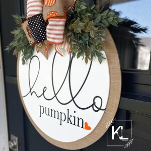Puede incluir: Un colgador de puerta redondo de madera con el texto "hello pumpkin" en escritura negra con un peque&ntilde;o coraz&oacute;n naranja. El colgador est&aacute; decorado con un lazo de lunares negros y blancos con rayas naranja y blancas y una guirnalda de hojas verdes.