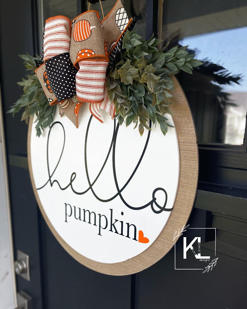 Puede incluir: Un colgador de puerta redondo de madera con el texto "hello pumpkin" en escritura negra con un peque&ntilde;o coraz&oacute;n naranja. El colgador est&aacute; decorado con un lazo de lunares negros y blancos con rayas naranja y blancas y una guirnalda de hojas verdes.