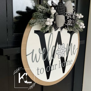 May include: A round wooden door hanger with a white background and a black letter "W" in the center. The word "welcome" is written in gray script above the letter and "to our home" is written in gray script below the letter. The letter "W" has a white snowflake in the center. The door hanger is decorated with a gray and black bow with white snowflakes and a sprig of green pine with white berries.
