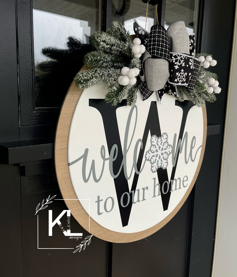 May include: A round wooden door hanger with a white background and a black letter "W" in the center. The word "welcome" is written in gray script above the letter and "to our home" is written in gray script below the letter. The letter "W" has a white snowflake in the center. The door hanger is decorated with a gray and black bow with white snowflakes and a sprig of green pine with white berries.