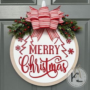 May include: A round wooden door sign with a white center and light wood border. It features red text reading MERRY CHRISTMAS, red snowflake cutouts, and two red outline pine trees. A large red and white striped bow sits atop with greenery.