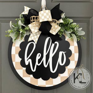 K&ouml;nnte beinhalten: Ein rundes T&uuml;rschild aus Holz mit einem schwarz-hellen Schachbrettmuster und einem zentralen schwarzen, gewellten Kreis, der den wei&szlig;en Schriftzug hello zeigt. Das Schild ist mit einer gro&szlig;en schwarz-wei&szlig;-beigen Schleife und gr&uuml;nem Blattwerk verziert.