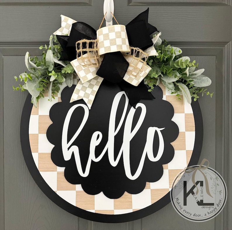 K&ouml;nnte beinhalten: Ein rundes T&uuml;rschild aus Holz mit einem schwarz-hellen Schachbrettmuster und einem zentralen schwarzen, gewellten Kreis, der den wei&szlig;en Schriftzug hello zeigt. Das Schild ist mit einer gro&szlig;en schwarz-wei&szlig;-beigen Schleife und gr&uuml;nem Blattwerk verziert.