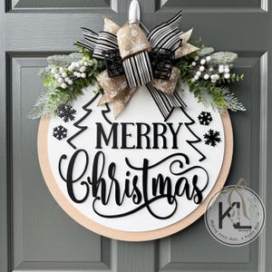 May include: A round wooden door sign with a white center and natural wood border. It features black cutout text reading Merry Christmas, black snowflake accents, and a large bow with beige and black striped ribbons and greenery.