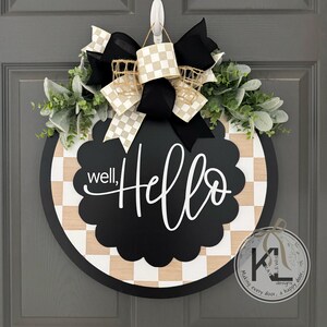 K&ouml;nnte beinhalten: Ein rundes T&uuml;rschild aus Holz mit einer schwarzen, gewellten Mitte und dem wei&szlig;en Schriftzug well, Hello. Der Hintergrund zeigt ein helles Holz- und wei&szlig;es Schachbrettmuster. Es ist mit einer gro&szlig;en schwarz-beigen Schleife und gr&uuml;nem Laub verziert.