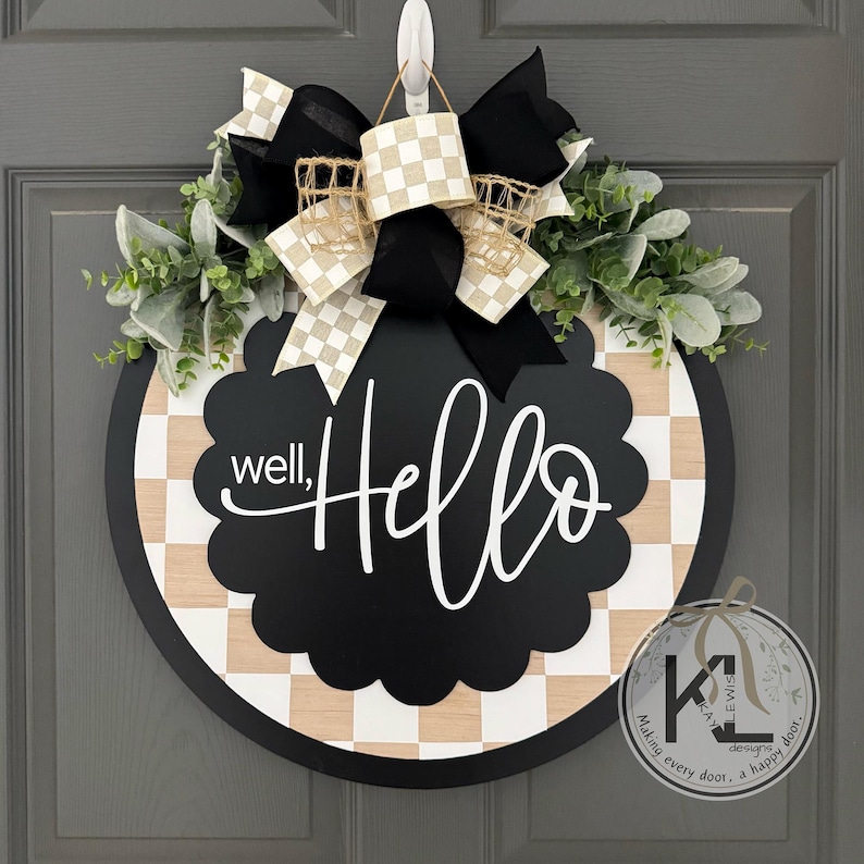 K&ouml;nnte beinhalten: Ein rundes T&uuml;rschild aus Holz mit einer schwarzen, gewellten Mitte und dem wei&szlig;en Schriftzug well, Hello. Der Hintergrund zeigt ein helles Holz- und wei&szlig;es Schachbrettmuster. Es ist mit einer gro&szlig;en schwarz-beigen Schleife und gr&uuml;nem Laub verziert.