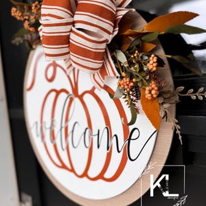 May include: A round wooden door hanger with a welcome sign and a pumpkin design. The pumpkin is orange and the welcome sign is in black script. The hanger is decorated with a brown and white striped bow and fall foliage.