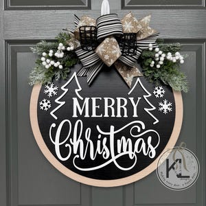 May include: A round black wooden door sign with the white text Merry Christmas, featuring white pine tree and snowflake cutouts. The top is decorated with a large bow in black and white stripes and tan snowflake patterns, plus white berries.