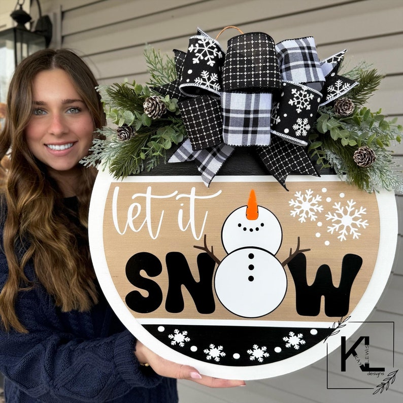 May include: A round wooden door hanger with a black and white plaid bow, evergreen sprigs, and pine cones. The door hanger features a snowman and the text "Let it Snow".
