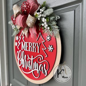 May include: A round red wooden door sign with the white text Merry Christmas, featuring two white pine tree silhouettes and snowflake cutouts. It is decorated with a large bow of red and white striped and tan snowflake ribbon, plus frosted greenery and white berries.