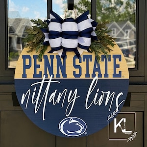 Puede incluir: Un colgador de puerta redondo de madera con un dise&ntilde;o azul y dorado de Penn State Nittany Lions. La parte superior del colgador de puerta tiene un lazo a rayas blanco y azul con hojas verdes.