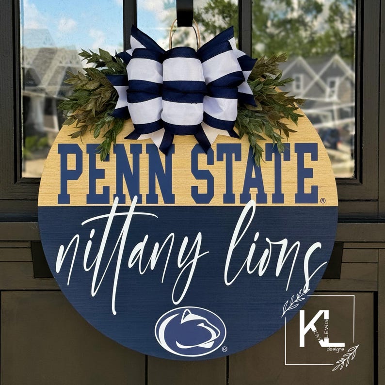 Puede incluir: Un colgador de puerta redondo de madera con un dise&ntilde;o azul y dorado de Penn State Nittany Lions. La parte superior del colgador de puerta tiene un lazo a rayas blanco y azul con hojas verdes.