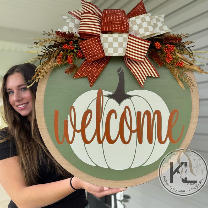 Pode incluir: Uma placa de boas-vindas redonda de madeira com um design de ab&oacute;bora branca e a palavra "welcome" em escrita laranja. A placa &eacute; decorada com um grande la&ccedil;o com padr&otilde;es xadrez e listrados vermelhos e creme, e folhagem de outono.