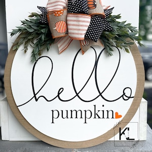 Puede incluir: Un letrero redondo de madera con fondo blanco y una inscripci&oacute;n cursiva negra que dice "hello pumpkin" con un peque&ntilde;o coraz&oacute;n naranja. El letrero est&aacute; decorado con un lazo hecho de tela estampada naranja, blanca y negra con un peque&ntilde;o dise&ntilde;o de calabaza. El lazo est&aacute; acentuado con ramitas verdes.