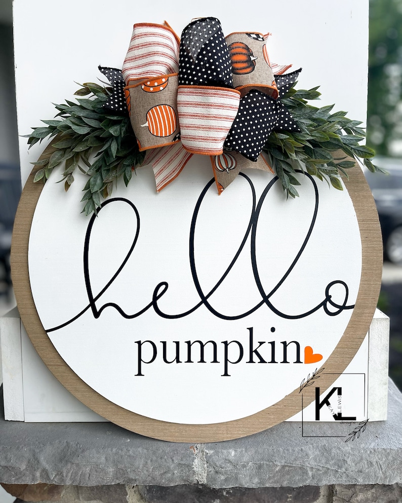 Puede incluir: Un letrero redondo de madera con fondo blanco y una inscripci&oacute;n cursiva negra que dice "hello pumpkin" con un peque&ntilde;o coraz&oacute;n naranja. El letrero est&aacute; decorado con un lazo hecho de tela estampada naranja, blanca y negra con un peque&ntilde;o dise&ntilde;o de calabaza. El lazo est&aacute; acentuado con ramitas verdes.