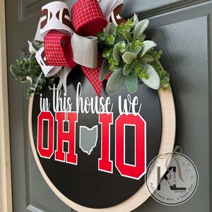 May include: A round black wooden door sign featuring the white text in this house we and large red block letters spelling OH-IO with a gray map of Ohio. The sign is decorated with a multi-layered bow featuring football patterns and green foliage.