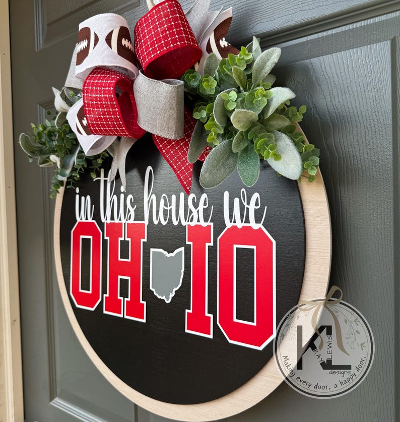 May include: A round black wooden door sign featuring the white text in this house we and large red block letters spelling OH-IO with a gray map of Ohio. The sign is decorated with a multi-layered bow featuring football patterns and green foliage.