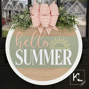 Pu&ograve; includere: Un appendiporta rotondo in legno con un bordo bianco. L'appendiporta ha uno sfondo in legno verde e marrone con il testo "hello summer" in lettere rosa e bianche. Un grafico del sole giallo &egrave; sopra il testo. Un fiocco a righe rosa e bianco con ramoscelli verdi &egrave; attaccato alla parte superiore dell'appendiporta.