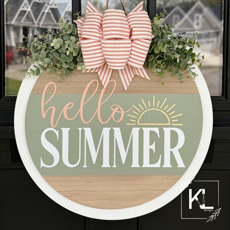 Pu&ograve; includere: Un appendiporta rotondo in legno con un bordo bianco. L'appendiporta ha uno sfondo in legno verde e marrone con il testo "hello summer" in lettere rosa e bianche. Un grafico del sole giallo &egrave; sopra il testo. Un fiocco a righe rosa e bianco con ramoscelli verdi &egrave; attaccato alla parte superiore dell'appendiporta.