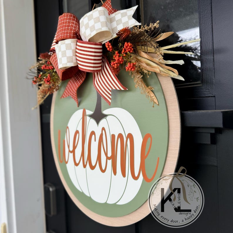 Pode incluir: Uma placa de boas-vindas redonda de madeira com um design de ab&oacute;bora. A placa apresenta um fundo verde com uma ab&oacute;bora branca e a palavra "welcome" em letras laranja. &Eacute; decorada com um la&ccedil;o e folhagem de outono.