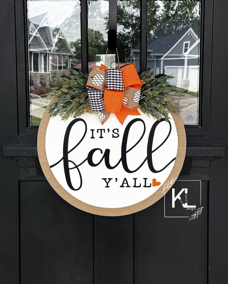 May include: A round, wooden door hanger with the text "It's fall y'all" in black script. The hanger is decorated with a wreath of green leaves and orange and black ribbon.