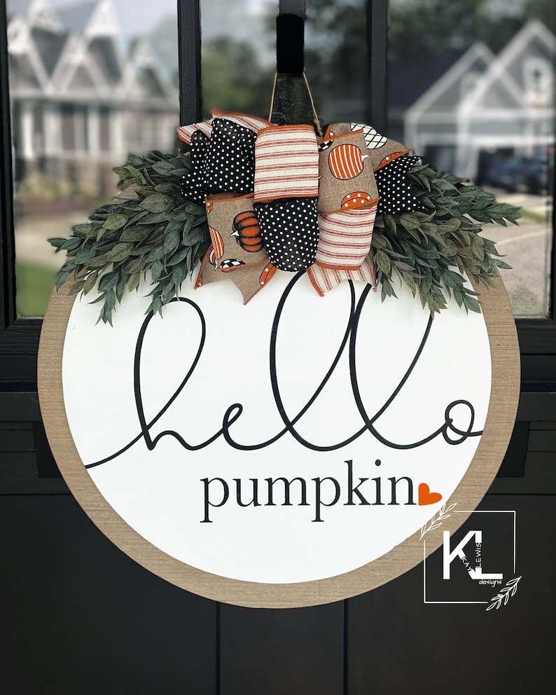 Puede incluir: Colgador de puerta redondo de madera con borde de arpillera. El letrero dice "hello pumpkin" con un coraz&oacute;n rojo. El letrero est&aacute; decorado con un gran lazo de tela con estampado naranja, blanco y negro.