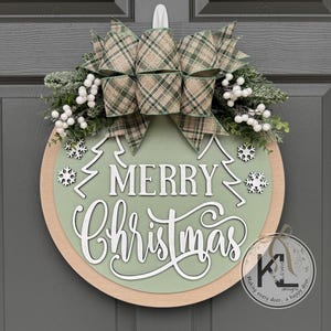 May include: A round wooden door sign featuring a sage green center with the white text Merry Christmas, two white pine tree outlines, and white snowflake cutouts. It is topped with a plaid bow, frosted greenery, and white berries.