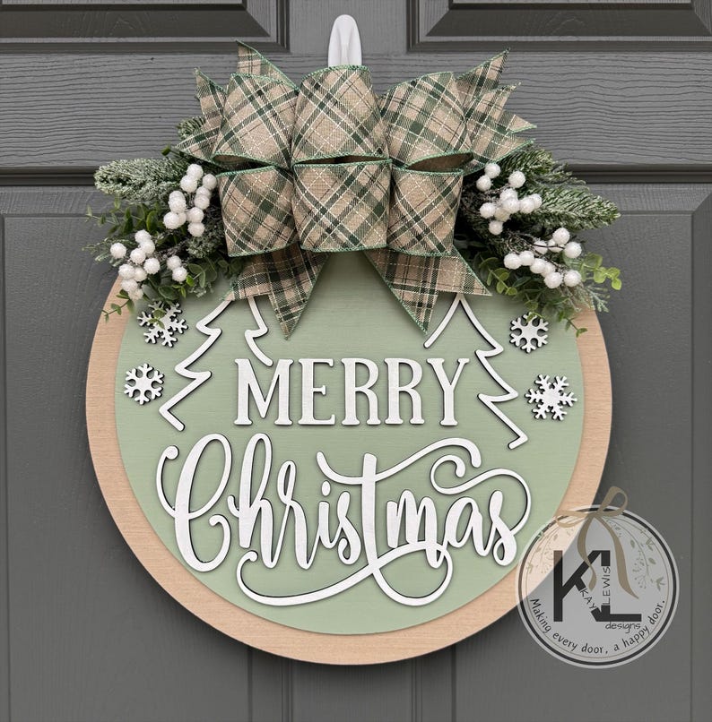 May include: A round wooden door sign featuring a sage green center with the white text Merry Christmas, two white pine tree outlines, and white snowflake cutouts. It is topped with a plaid bow, frosted greenery, and white berries.