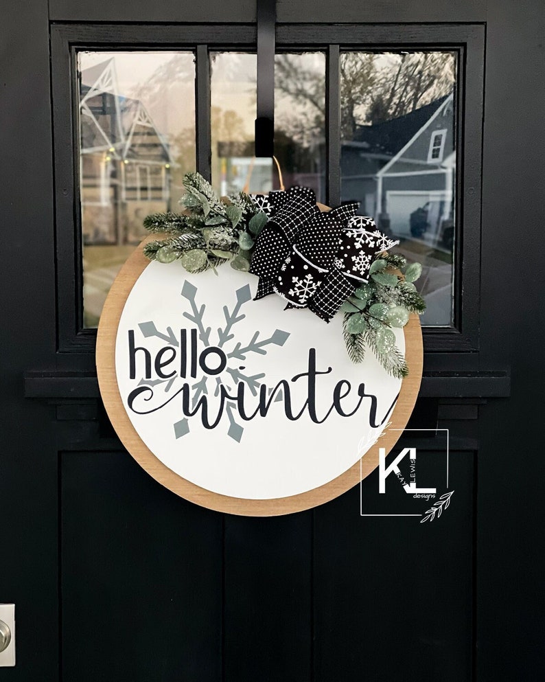 May include: A round wooden door hanger with a white background and the text "hello winter" in black script. A large gray snowflake is behind the text. The door hanger is decorated with a black and white polka dot bow and green sprigs.