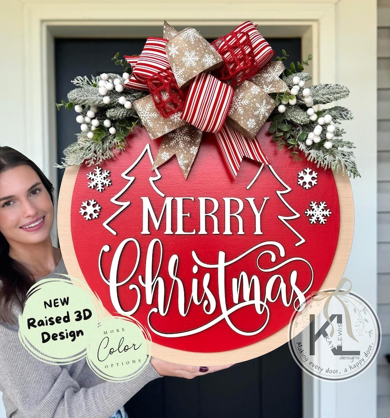 May include: A round wooden door sign with a red center featuring the white text Merry Christmas and two white tree outlines. It is decorated with a large bow of red and white striped and tan snowflake-patterned ribbon, plus frosted greenery and white berries.