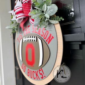 May include: A wooden door wreath with a football design. The wreath features the text "'TIS THE SEASON GO BUCKS!" in red. The football has a red "O" in the centre. The wreath is decorated with greenery and a red and white bow.