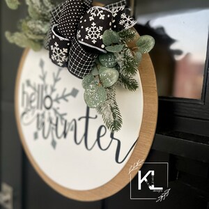 May include: A white round wooden sign with the text "hello winter" in black script. The sign is decorated with a black and white polka dot and snowflake ribbon bow and a sprig of green eucalyptus.