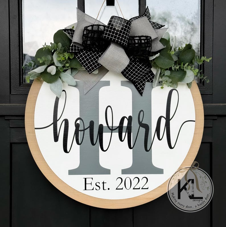 May include: A round wooden door hanger with a white background and a gray and black bow. The name "Howard" is written in black cursive lettering with the letters "H" in gray. The text "Est. 2022" is written in black below the name.