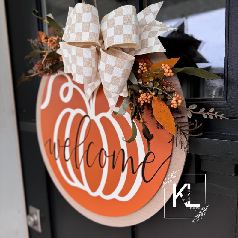 May include: A round wooden door hanger with a painted orange pumpkin design and the word "welcome" in black script. The pumpkin has a white outline and a stem. The door hanger is decorated with a beige and white checkered bow and a sprig of fall foliage with orange berries.