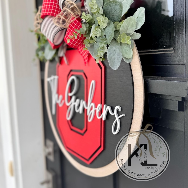 K&ouml;nnte beinhalten: Ein runder Holzt&uuml;rh&auml;nger mit einem rot-schwarzen Logo der Ohio State University und dem Namen "The Gerbers" in wei&szlig;er Schrift. Der T&uuml;rh&auml;nger ist mit einer rot-wei&szlig; gestreiften Schleife und Gr&uuml;npflanzen verziert.