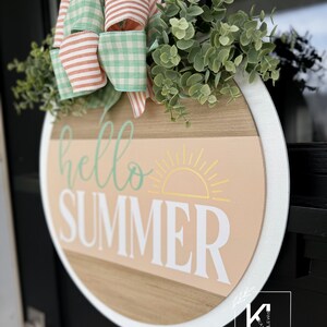 Pu&ograve; includere: Un appendiporta rotondo in legno con un bordo bianco. L'appendiporta ha uno sfondo pesca con un sole giallo e le parole "hello summer" in lettere bianche e verdi. Un fiocco di Vichy verde e bianco e un ramoscello di verde sono attaccati alla parte superiore dell'appendiporta.